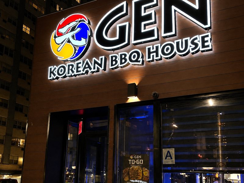 The lit GEN Korean BBQ House storefront at night, with the entrance and dining room windows below the sign.